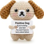 A grey crochet dog miniature figure with floppy ears and blush details, holding a sign with an encouraging message.