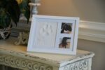 White wooden picture frame with a clay paw print impression and two photo slots, with a stencil kit beside it.