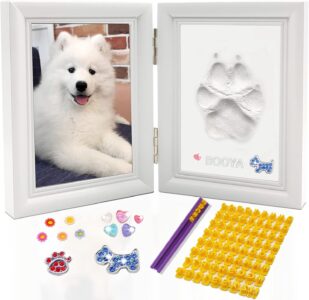A white rectangular wooden frame with a pet's paw print impression in clay, a photo slot, and decorative trinkets.