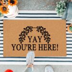 A colorful rectangular doormat with the humorous text "Yay You're Here!" in bold letters, set against a bright background.