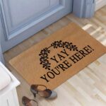 A colorful rectangular doormat with the humorous text "Yay You're Here!" in bold letters, set against a bright background.
