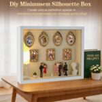 White XINGZUAN Museum of Us DIY Shadow Box Kit on a tabletop, displaying mini gold frames, figurines, and an LED light.